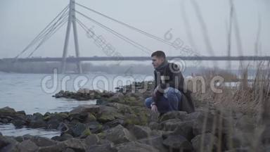 体贴的白人年轻人坐在河岸上向水中<strong>扔石头</strong>。 一幅画中悲伤的帅哥